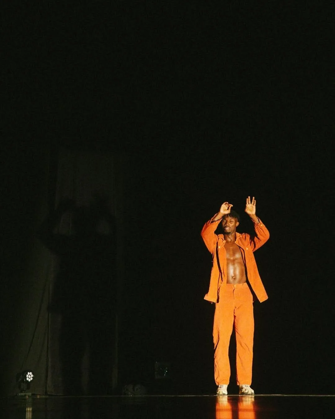 Dancer in orange on a dark stage raising his hands.