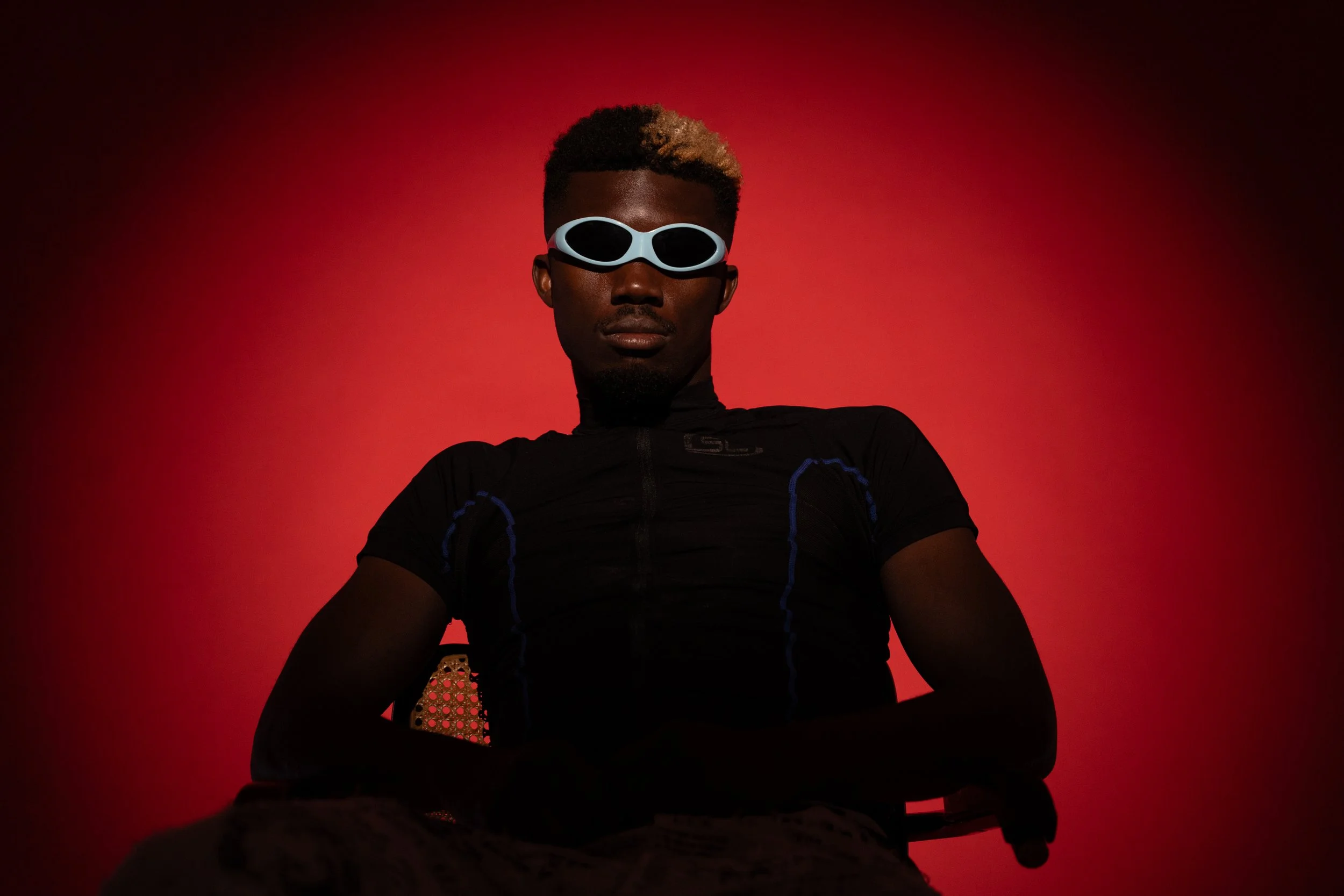 Oulouy wearing a black athletic shirt and white futuristic sunglasses against a red background.
